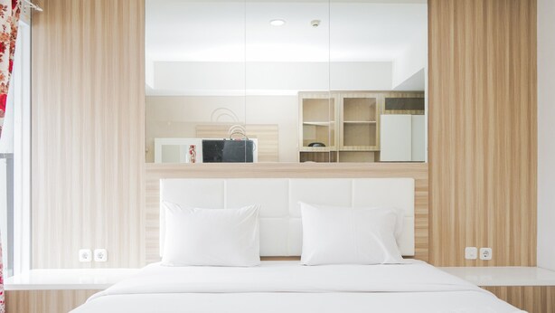 A modern hotel room with a large bed, white bedding, a wooden headboard, a built-in desk, and a mirror above the bed. - Simply Strategic and Comfy Studio Paramount Skyline Apartment (Gading Serpong)