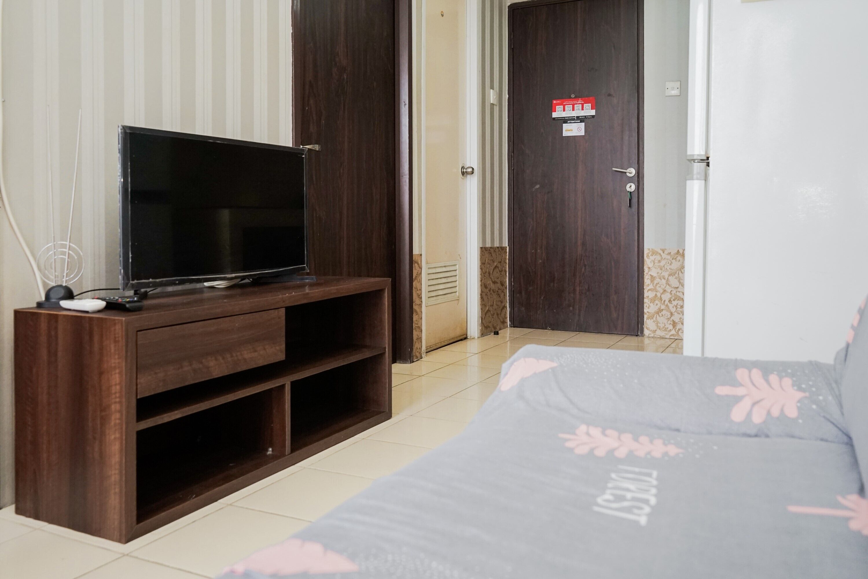 Une chambre d’hôtel avec un meuble en bois pour la télévision, un lit recouvert d’une couvre-lit à motifs floraux et une porte sur laquelle est apposé un panneau rouge indiquant la sortie de secours.