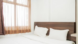 Un lit bien fait, avec du linge de lit blanc et deux oreillers, une tête de lit en bois et une fenêtre ornée de rideaux légers.