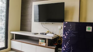 A room with a mounted TV on a wall, a small shelf with a box labeled "FIRST", and a refrigerator with a floral design. - Homey and Enjoy 2BR at Meikarta Apartment (Cikarang)