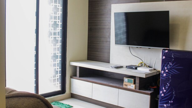 A modern living room with a flat-screen TV mounted on a wooden wall, a sofa, and a sliding glass door with a geometric pattern. - Homey and Enjoy 2BR at Meikarta Apartment (Cikarang)