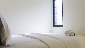 A neatly made bed with a white comforter and pillows, a window with a view of buildings, and two rolled towels on the bed. - Homey and Enjoy 2BR at Meikarta Apartment (Cikarang)