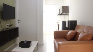 A living room with a brown leather sofa, a flat-screen TV mounted on the wall, a coffee table, and a door leading to another room.