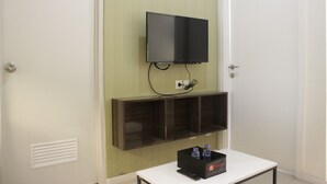 A room with a mounted flat-screen TV, a white table with a remote control, and a black box with bottled water.