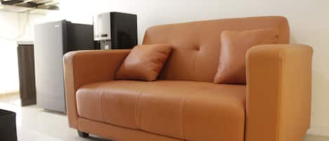 A tan leather sofa with two cushions, a white coffee table, and a black speaker in a modern room with white walls and a tiled floor.