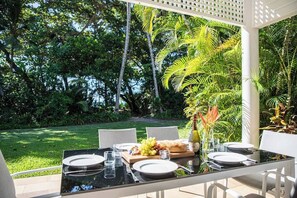Un área de comedor al aire libre con una mesa preparada para dos, rodeada de exuberante follaje y una pérgola blanca.