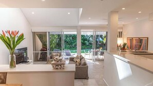 A modern living room with a sofa, a coffee table, and a TV. There is a glass door leading to an outdoor area with a lawn and chairs.