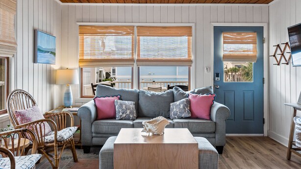 A cozy living room with a grey sofa, wooden chairs, and a blue door. - Adorable Ocean View 3 Bed/1 Bath Cottage Sleeps 8 steps to the beach (Ocean Isle Beach)
