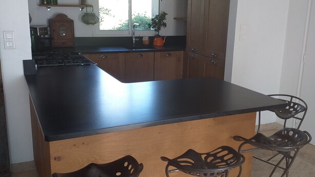 A kitchen with a black countertop, wooden cabinets, and a window. There are two metal chairs with holes in the seats. - Maison à la Campagne 15min de la mer (Beaulieu-Sous-la-Roche)