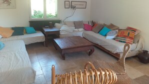 A cozy living room with a sofa, a wooden coffee table, and a wicker chair. There is a window with blinds, a radiator, and a lamp on a table. - Maison à la Campagne 15min de la mer (Beaulieu-Sous-la-Roche)