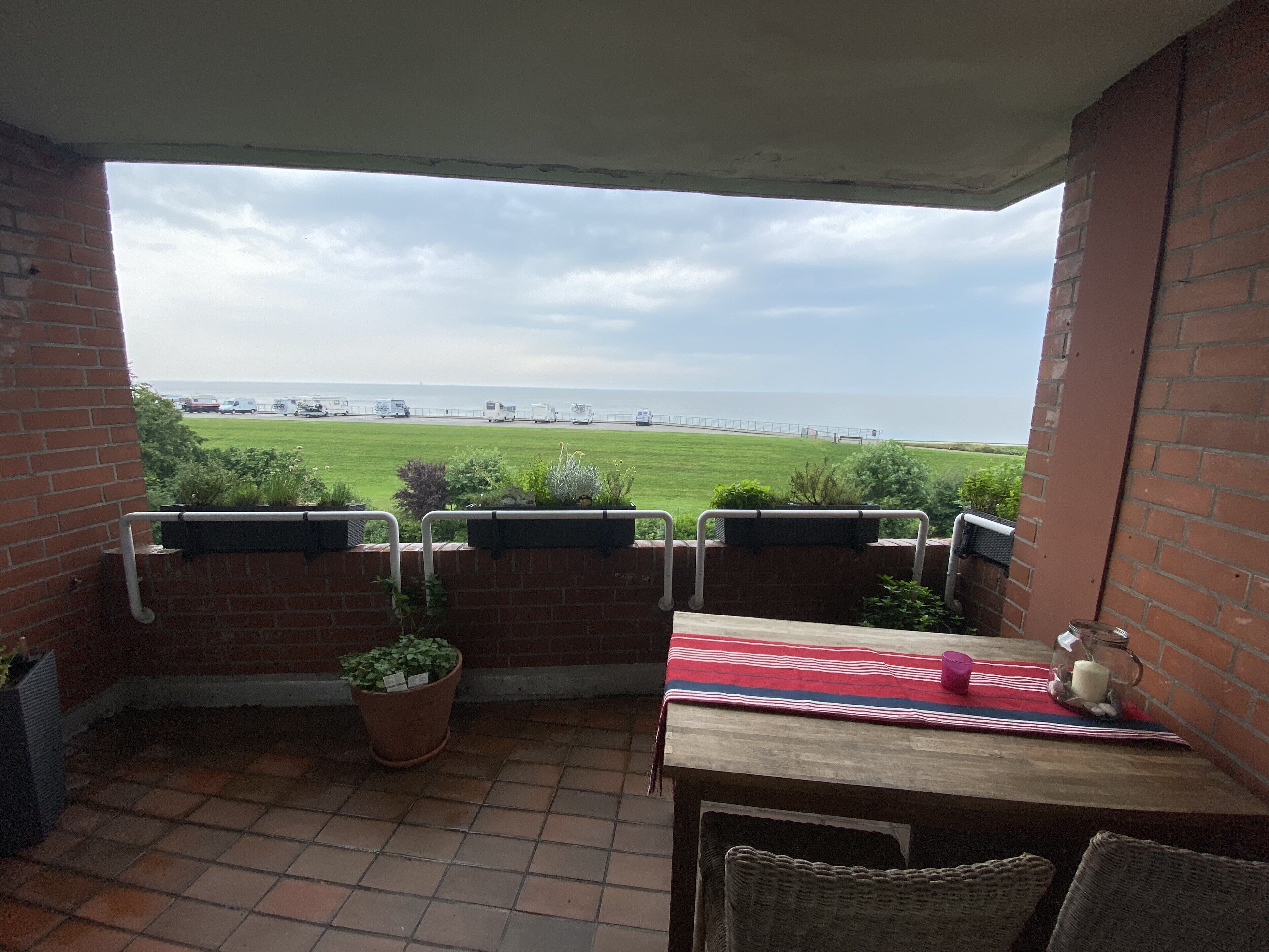 Une terrasse avec vue sur la mer, un mur de briques, une table recouverte d’une nappe à rayures rouges et blanches, et des plantes en pot.