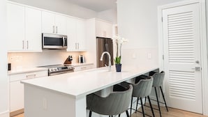 Une cuisine moderne avec des meubles blancs, un réfrigérateur en acier inoxydable, un four à micro-ondes et une cuisinière à gaz. On y trouve quatre tabourets de bar gris et une orchidée en pot sur l’îlot central.