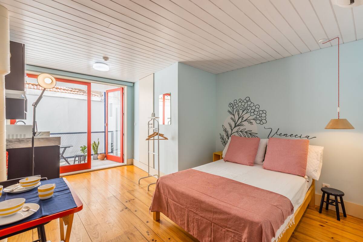 A bedroom with a bed, a desk with a lamp, a hanging plant, and a balcony with a table and chairs.