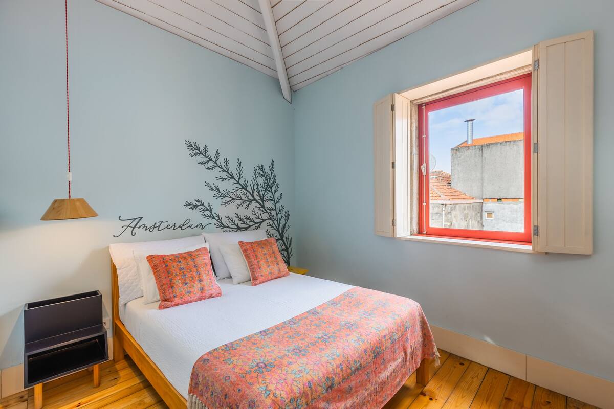 A bedroom with a bed, a window, a wall-mounted artwork, and a wooden floor.