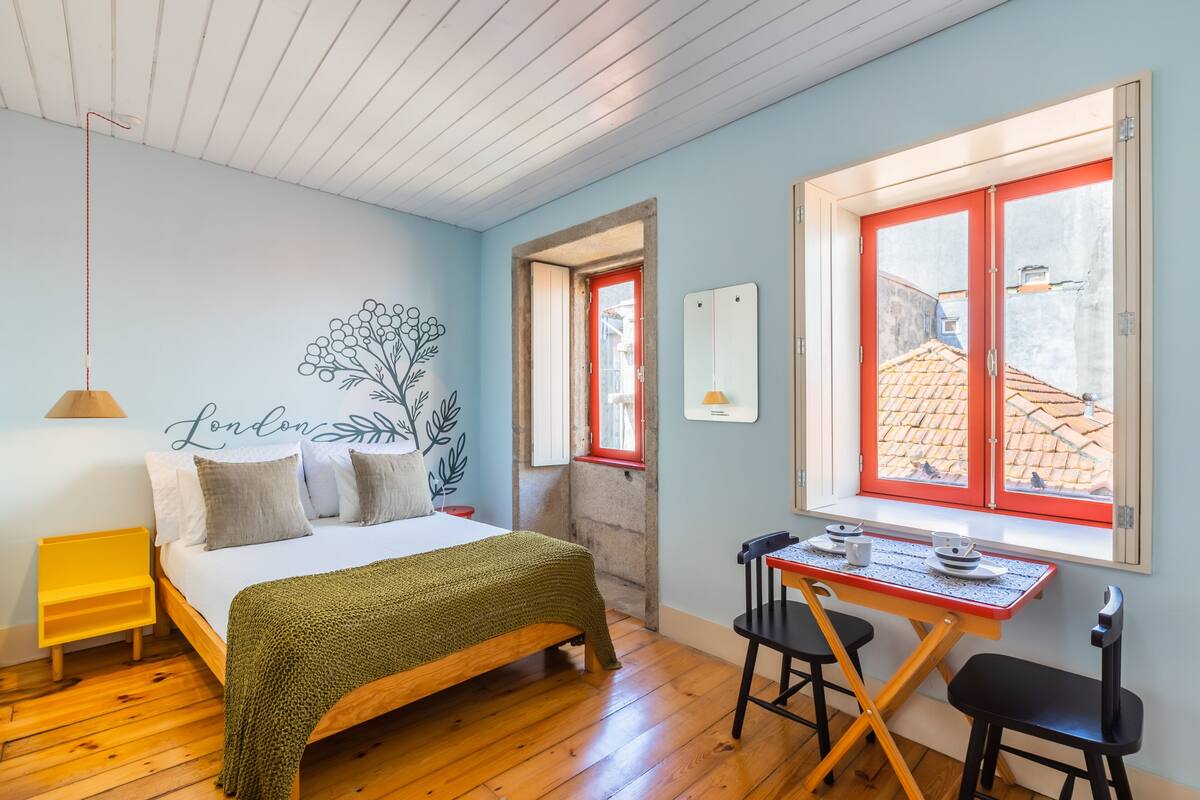 A bedroom with a bed, a desk, and a chair. There is a window with red frames and a view of buildings outside.