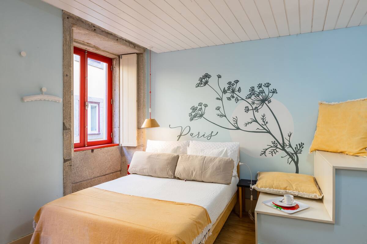 A bedroom with a bed, a window with a red frame, a wall with a floral mural, and a small table with a cup and saucer.