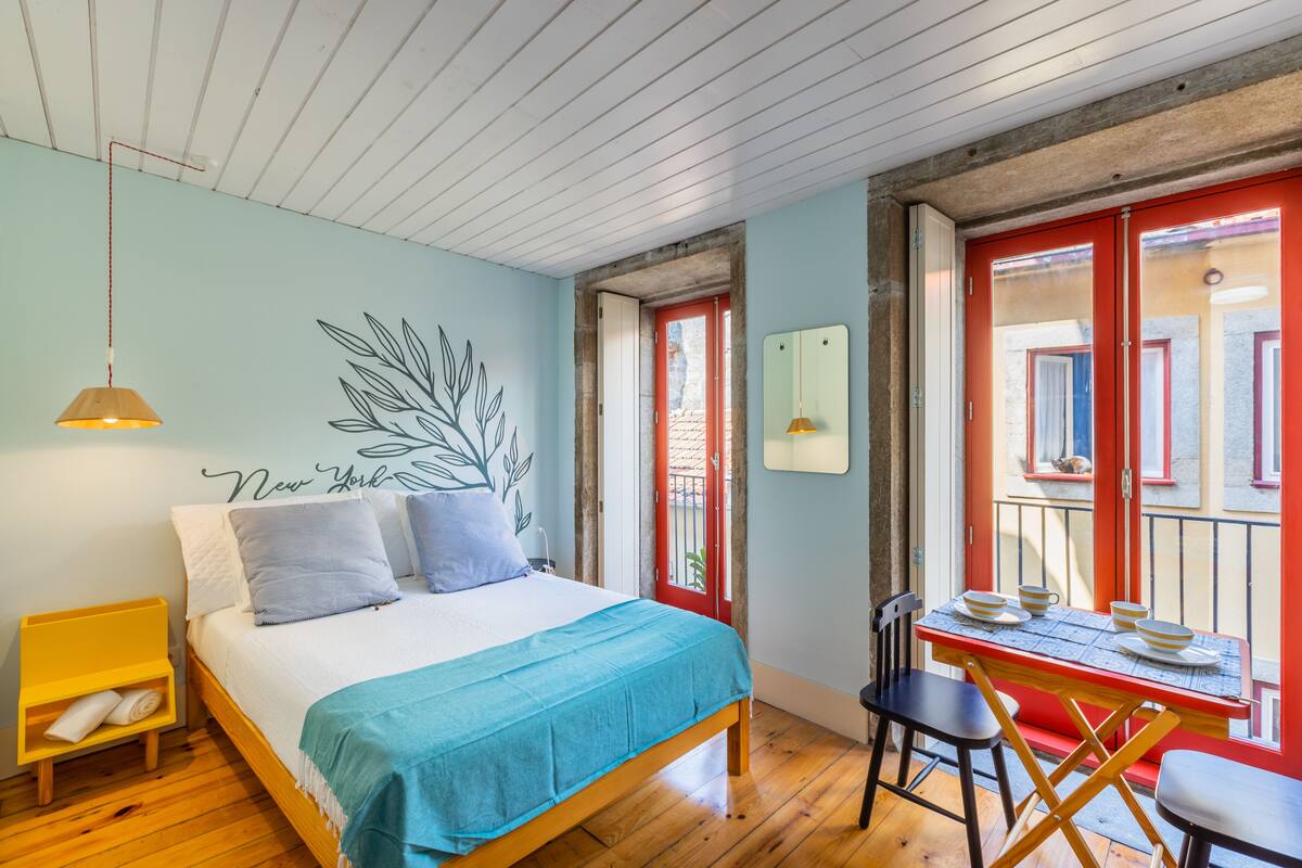 A bedroom with a bed, a table with chairs, a hanging lamp, and a red door.