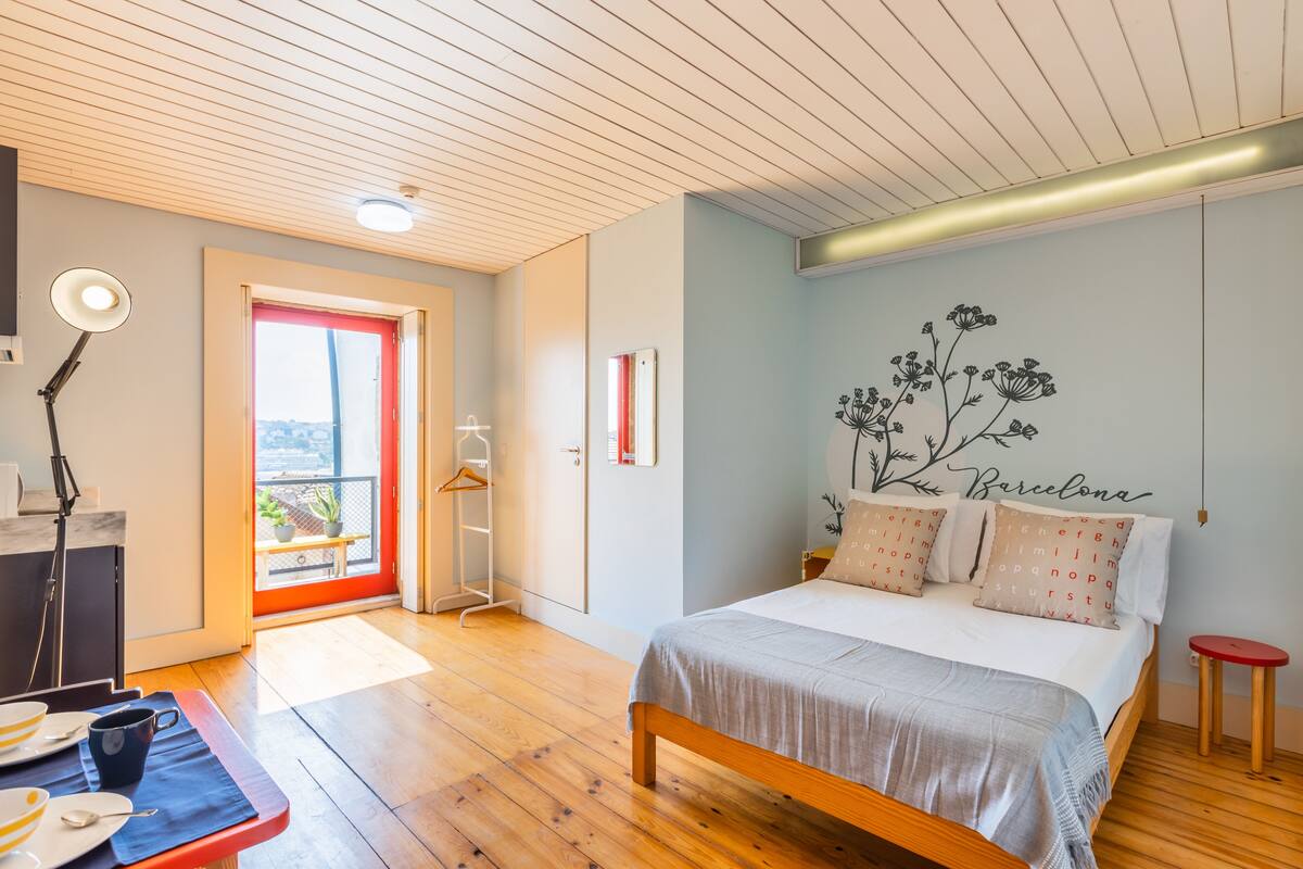 A bedroom with a bed, a desk with a lamp, a red door, and a balcony with a view.