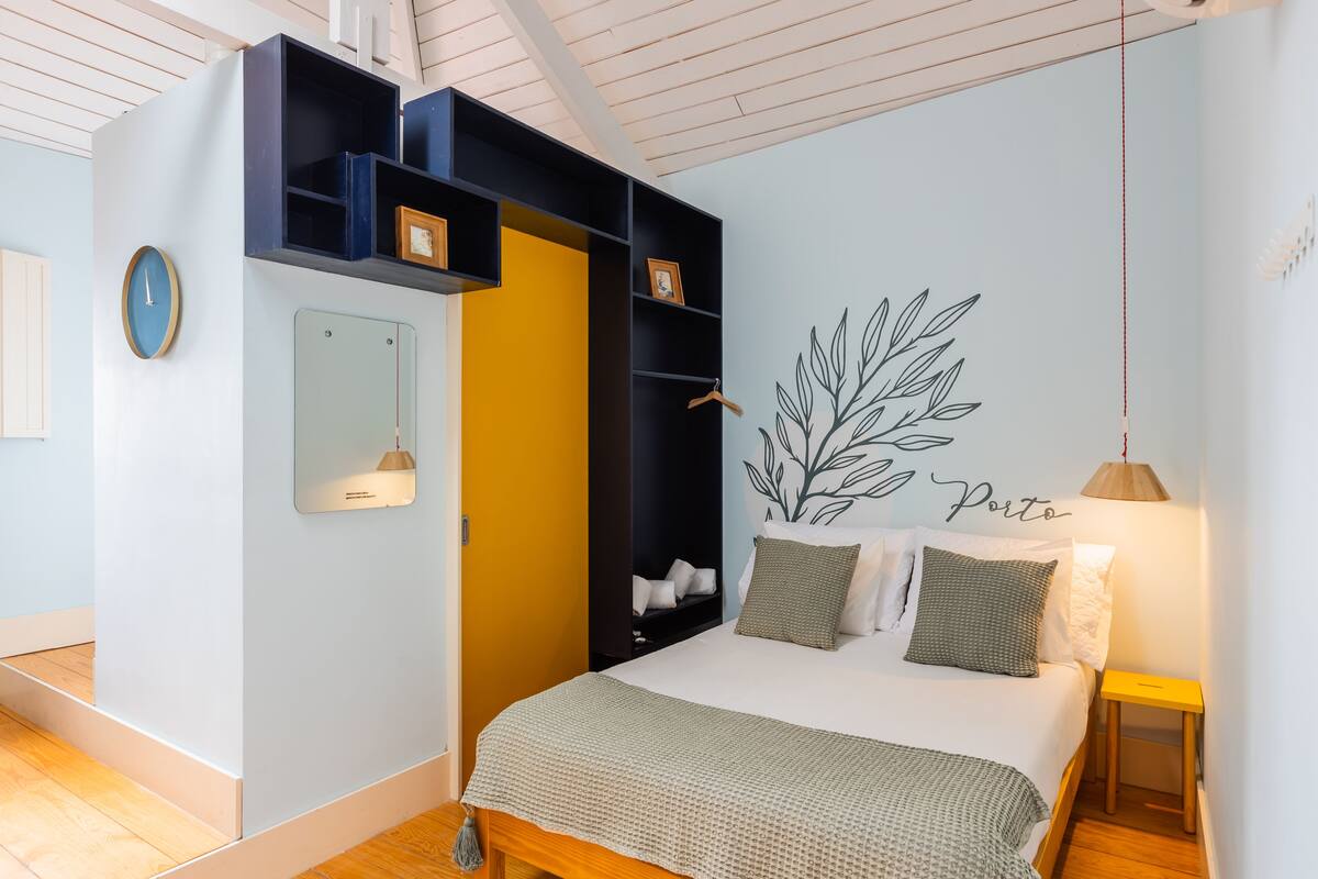 A bedroom with a bed, a wardrobe, a mirror, a wall clock, and a wall-mounted plant.
