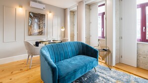 A modern living room with a blue sofa, a dining table, and a chair. There is a large window with curtains, a wall-mounted air conditioner, and a painting on the wall. - Santa Catarina Prime Studios (Porto)