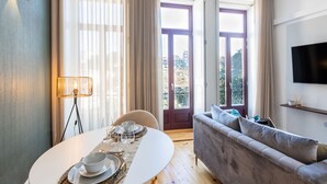 A modern living room with a round dining table set for two, a sofa, a TV mounted on the wall, and a balcony with a view of buildings. - Santa Catarina Prime Studios (Porto)