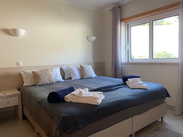 A neatly made bed with pillows and folded towels, a bedside table, and a window with curtains.