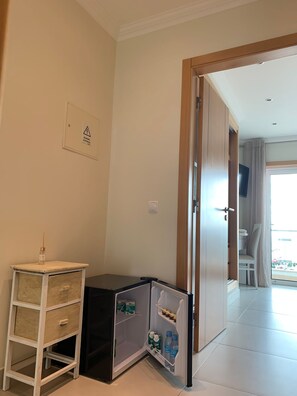 A compact room with a small refrigerator, a bedside table, and a door leading to a balcony.