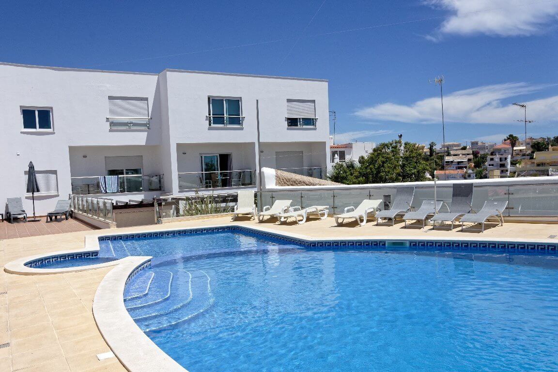 Una piscina con sillas de descanso, un edificio blanco con varias ventanas y un cielo despejado.