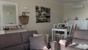 A living room with a sofa, a dining table with chairs, a wall-mounted air conditioner, and a decorative wall piece. - Apartment Oasis Praia Carvoeiro Bay (Lagoa)