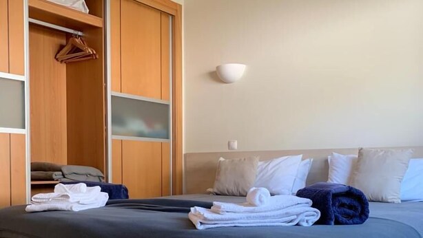 A neatly made bed with folded towels, a wooden wardrobe with a mirror, and a wall-mounted light fixture. - Apartment Oasis Praia Carvoeiro Bay (Lagoa)
