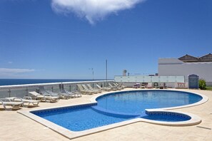 A swimming pool with lounge chairs, a view of the ocean, and a building with a flat roof.