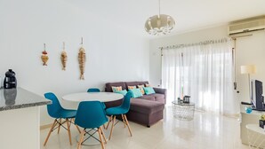 A modern living room with a round dining table, blue chairs, a sofa with cushions, a hanging light fixture, and a TV on a stand. - Quinta das Palmeiras Pool & Beach in Pêra (Lagoa)