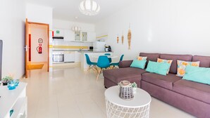 A modern living room with a brown sofa, teal cushions, and a white coffee table. A kitchen with white cabinets and blue chairs is visible in the background. - Quinta das Palmeiras Pool & Beach in Pêra (Lagoa)