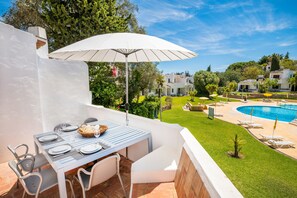 Uma área de refeições com uma mesa posta para quatro pessoas, um guarda-sol branco e vista para uma piscina e vegetação.