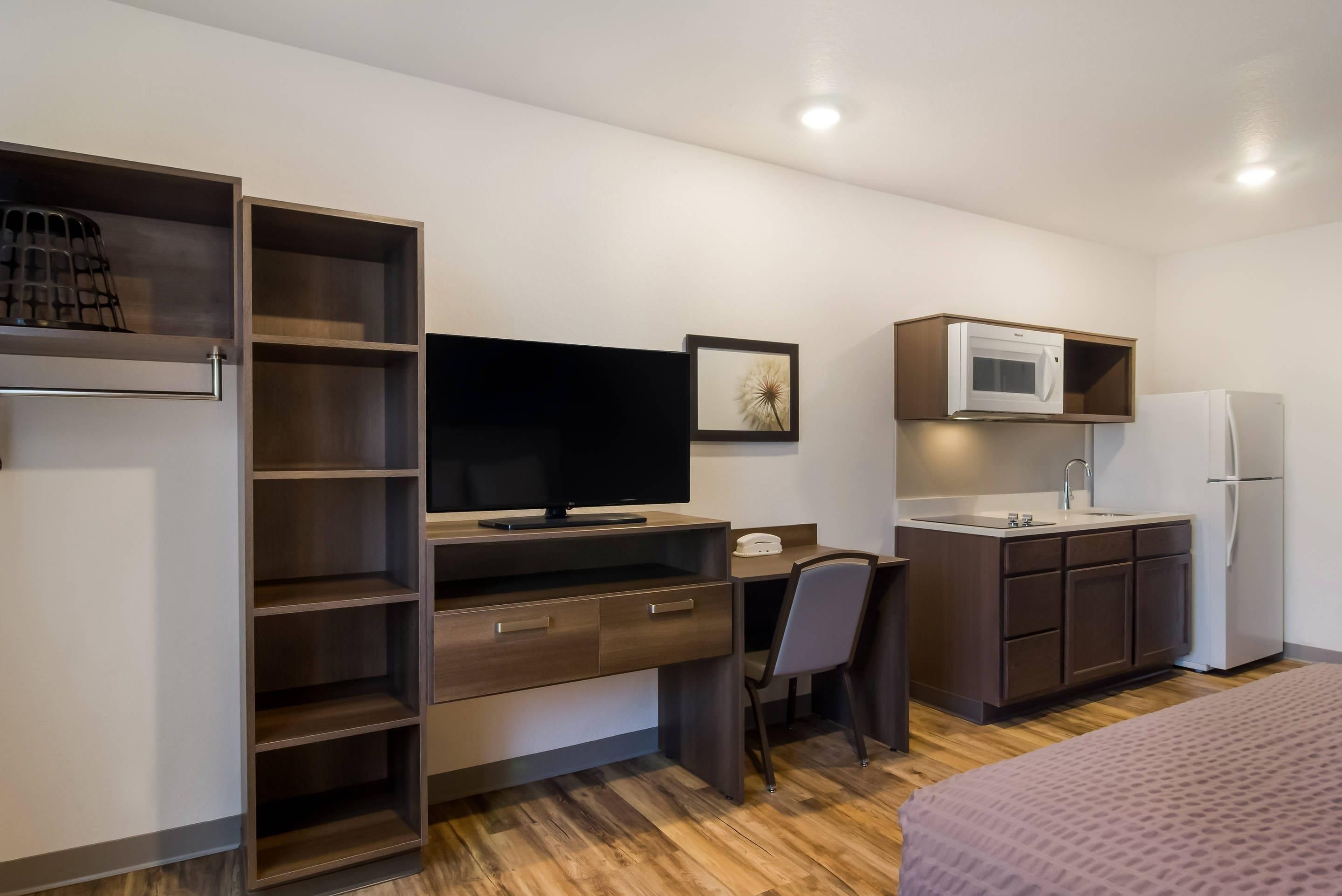 Habitación de hotel pequeña con kitchenette, un escritorio, un televisor de pantalla plana, una silla y una cama.