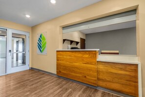 A reception area with a wooden reception desk, a glass door leading to a room, and a wall-mounted green leaf decoration. - WoodSpring Suites Jacksonville - South (Jacksonville)