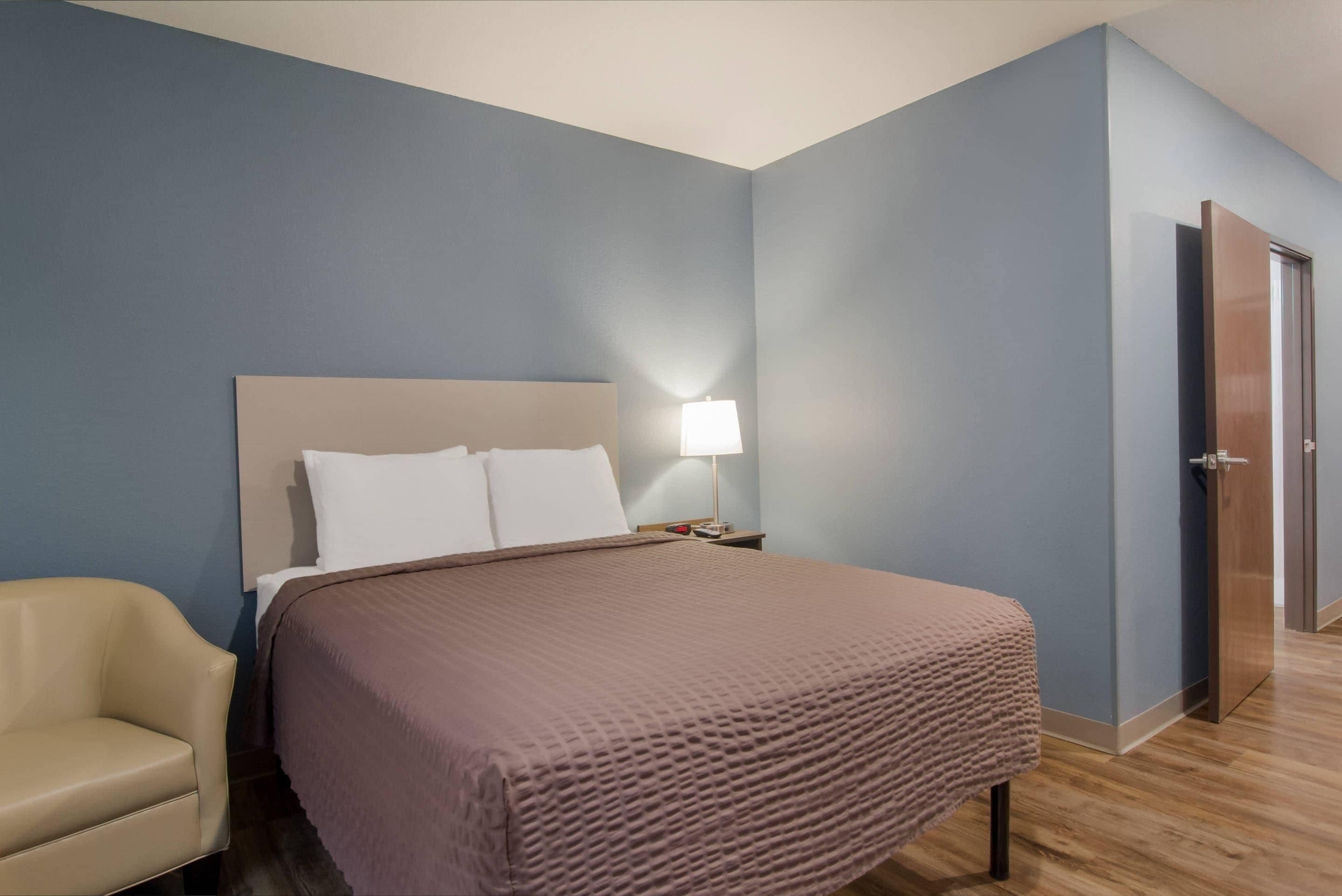 Una habitación de hotel con una cama, una silla, una mesita de noche, una lámpara y una puerta.