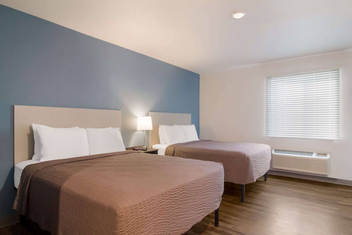 Une chambre d’hôtel avec deux lits, une table de chevet avec une lampe, une fenêtre avec des stores et un radiateur mural.