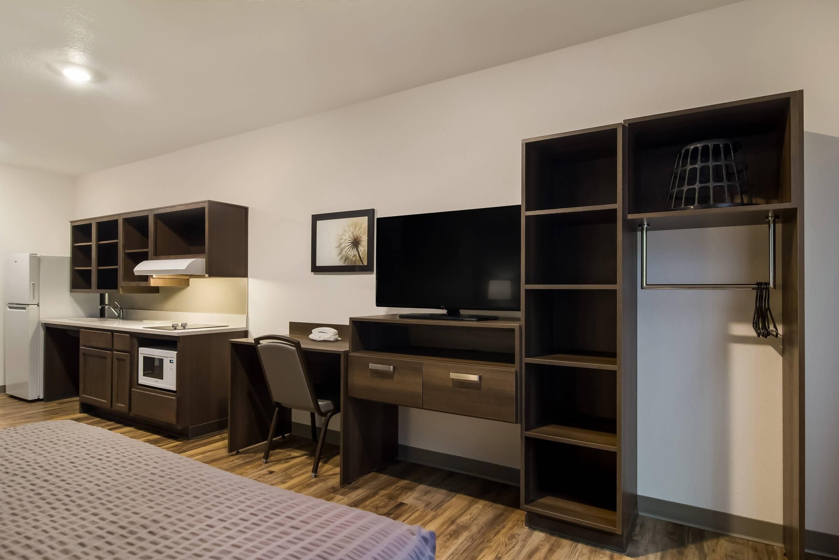 A compact hotel room with a kitchenette, a bed, a desk with a chair, a television, and a wardrobe.