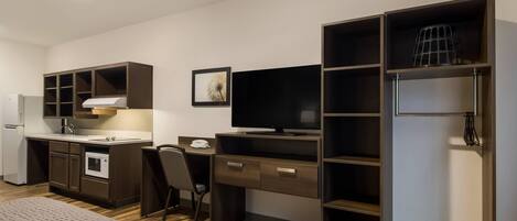 A compact hotel room with a kitchenette, a bed, a desk with a chair, a television, and a wardrobe.