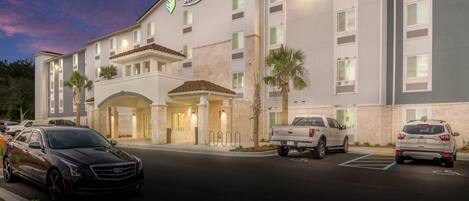 A hotel with a sign reading 'Woodspring Suites' and a parking lot with several vehicles.