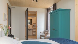 Ein Schlafzimmer mit einem Bett, einem türkisblauen Kleiderschrank, einer Holzbalkendecke und einem durch eine geöffnete Tür einsehbaren Badezimmer.