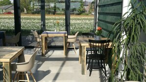 Eine Terrasse mit Holztischen und -stühlen, eine Glaswand mit Blick auf ein Feld und Häuser sowie eine grüne Wand mit Pflanzen.