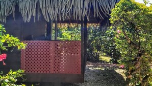Estructura con techo de paja y paneles celosía, rodeada de follaje y un sendero de grava.
