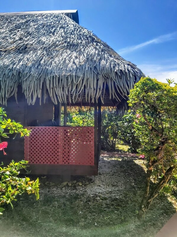 Estructura con techo de paja y paneles celosía, rodeada de follaje y un sendero de grava.