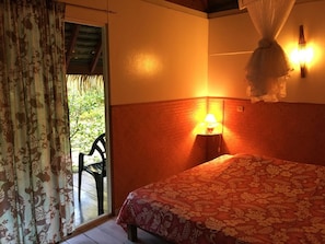 A room with a bed, a nightstand, a lamp, a chair, and a view of greenery outside.