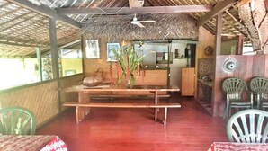Un restaurante con mesa y bancos de madera, techo de paja y sillas de plástico.