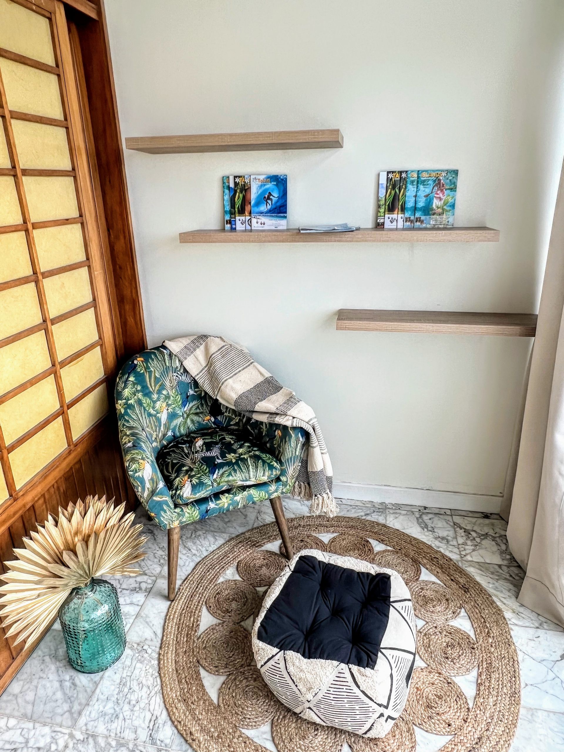 Une pièce d’angle dotée d’un paravent japonais traditionnel en papier de riz, d’un fauteuil à motifs, d’un tapis tissé, d’un petit coussin rond et de deux étagères murales où sont posés des livres.