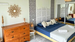 Une chambre à coucher avec une commode en bois, un lit avec une literie bleue, une petite table ronde et un grand miroir.