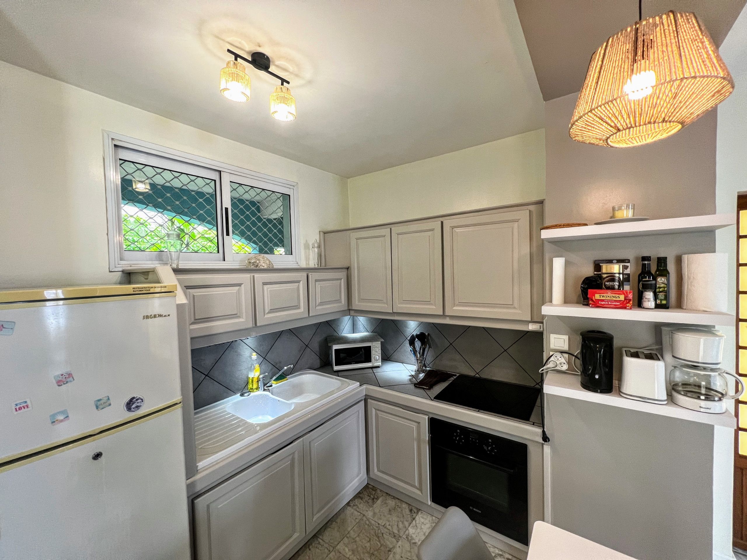 Une cuisine de taille réduite avec des meubles blancs, un réfrigérateur, un four à micro-ondes, un évier et une cuisinière. Il y a une fenêtre au-dessus de l’évier et un luminaire suspendu.